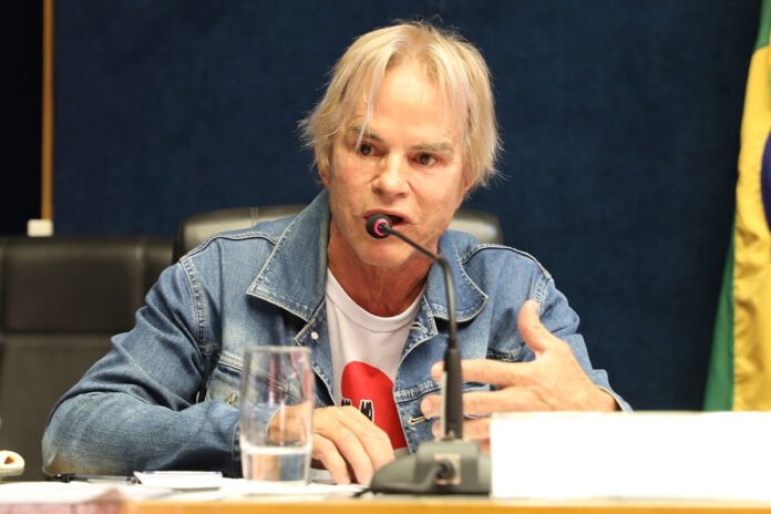 Homem de cabelo loiro e jaqueta jeans sentado ao microfone
