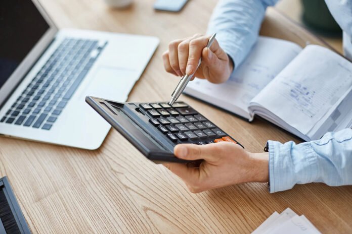 Homem manuseia calculadora; sobre a mesa estão relatórios